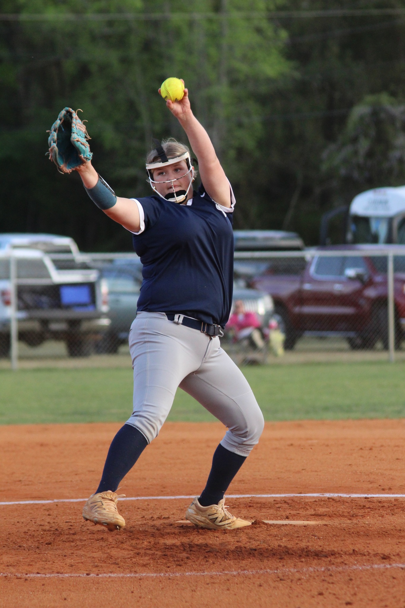 Softball pitcher in mid-throw, wearing a navy jersey and gray pants, standing on a dirt field with cars and trees in the background.