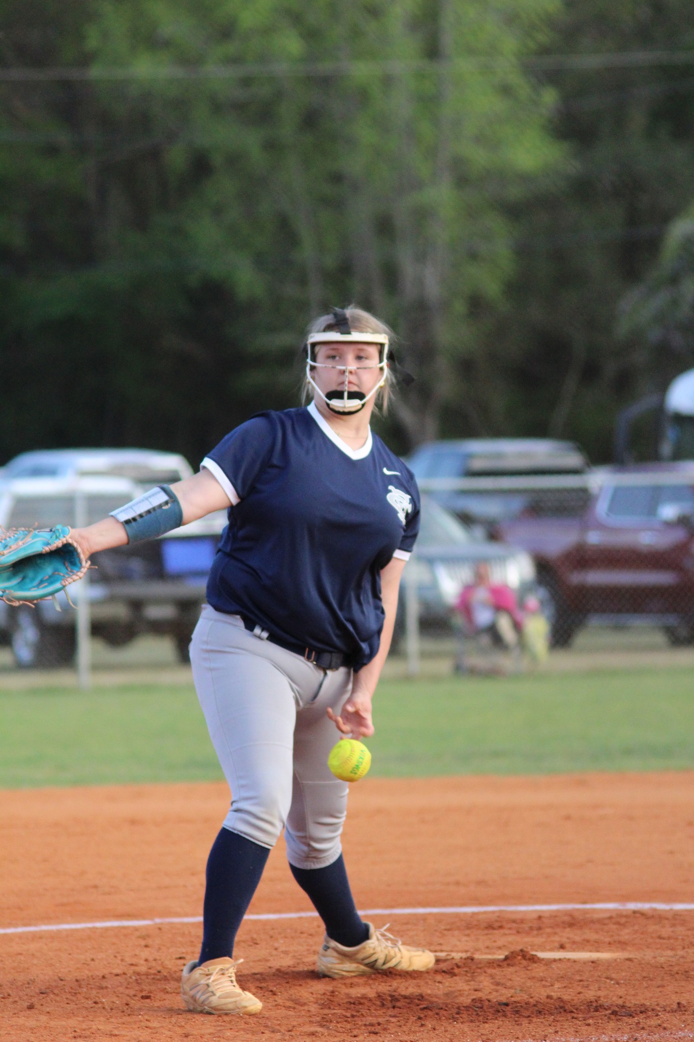 A softball player in navy jersey and gray pants prepares to pitch on a field, wearing a face guard, with cars and trees in the background.