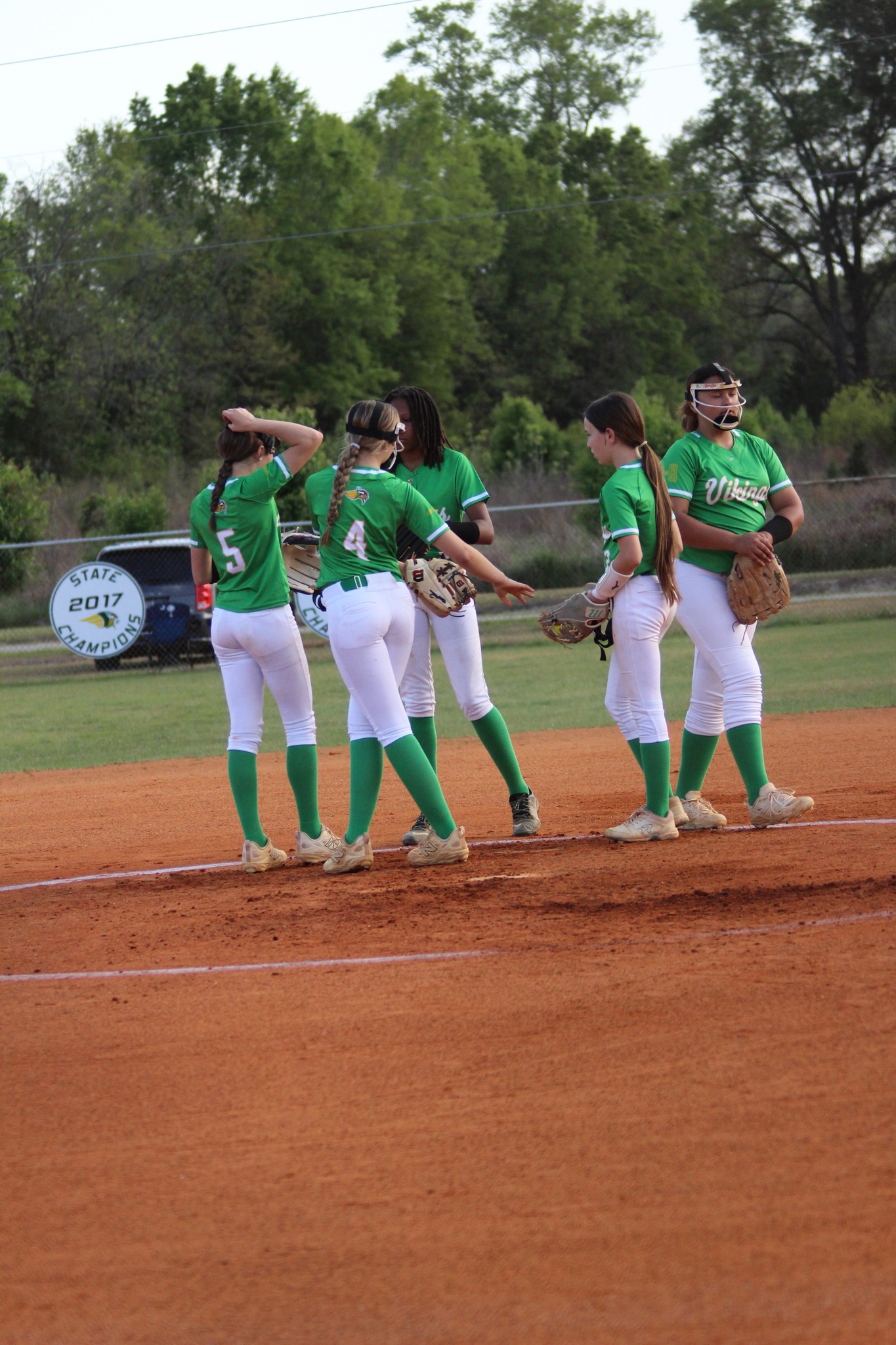 Four female softball players in green jerseys and white pants gather on a red dirt field, preparing for the game with trees and a fence in the background.