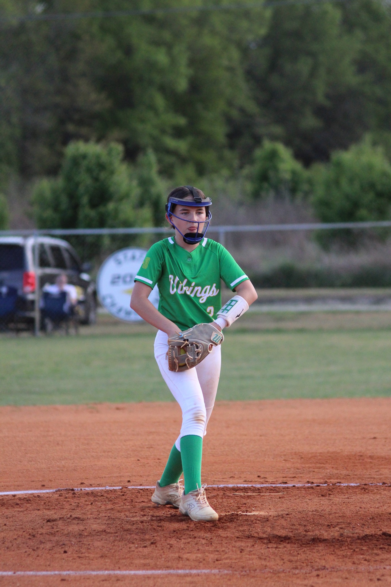 A softball player in a green jersey and white pants stands on a dirt field, wearing a helmet and glove, ready to pitch. Trees and a car are in the background.