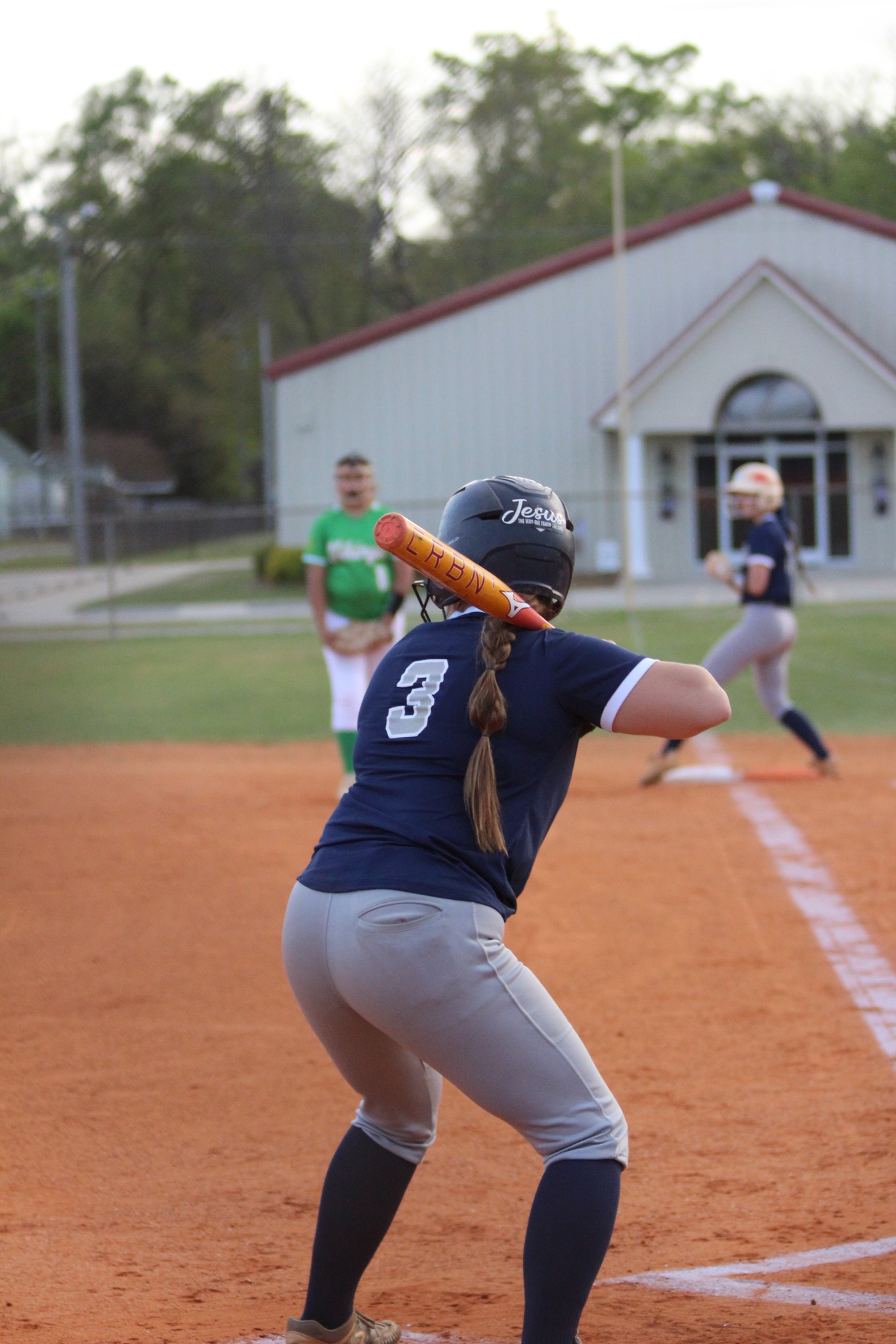 A softball player wearing a helmet and number 3 jersey prepares to bat, while other players and a building are visible in the background.