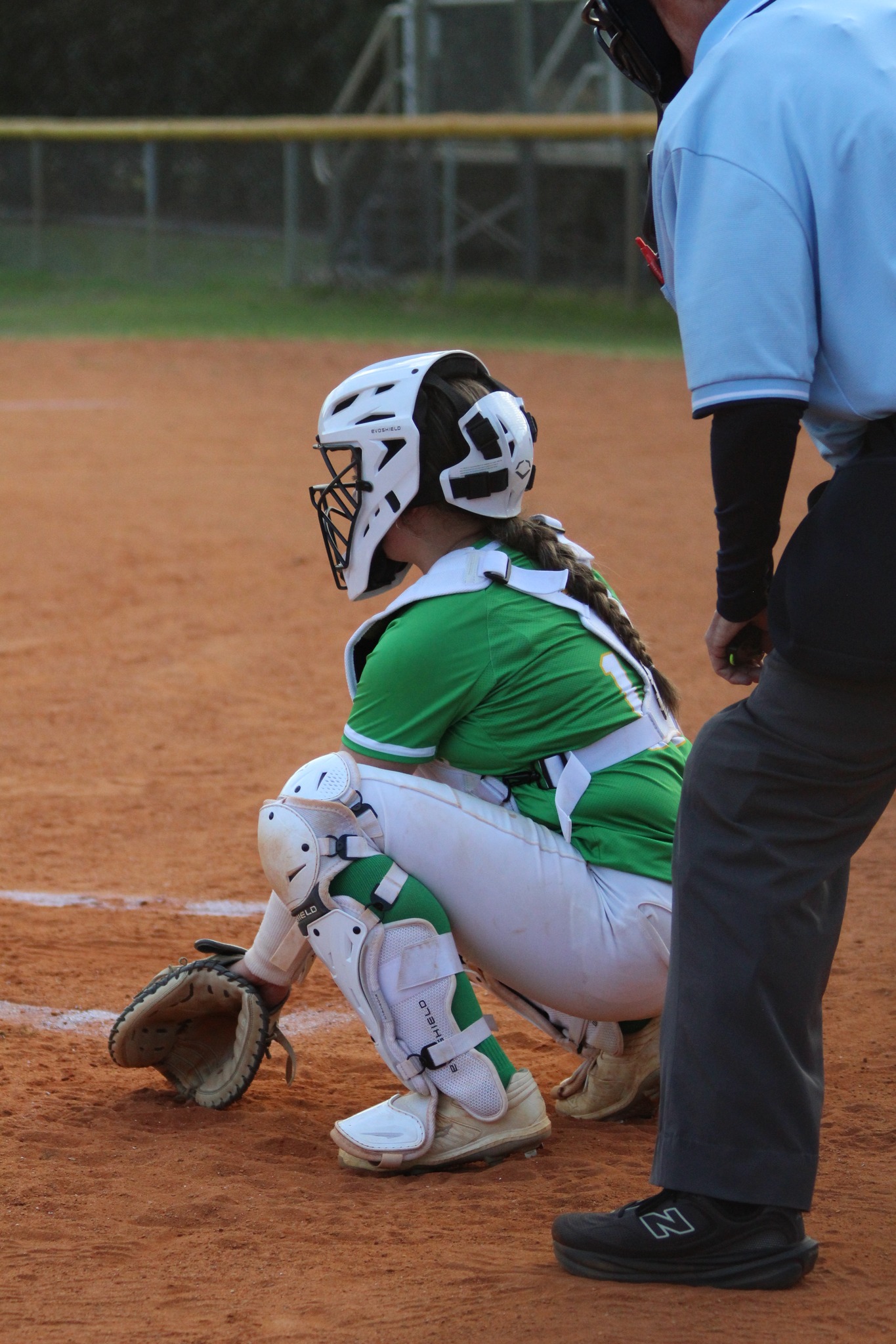 A softball catcher in green and white gear is crouched behind home plate, with an umpire standing nearby on a dirt field.