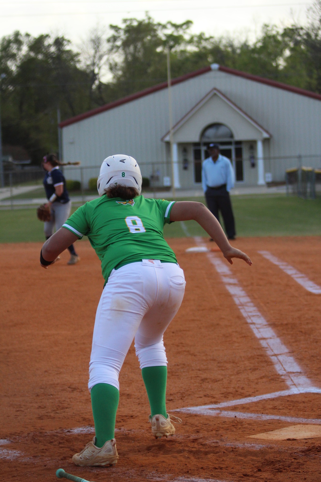 A softball player wearing a green jersey and white helmet runs toward a base on a dirt field, with an umpire and building in the background.
