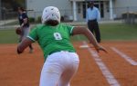 Softball player in green uniform running towards first base, with umpire and coach in background on a dirt field.