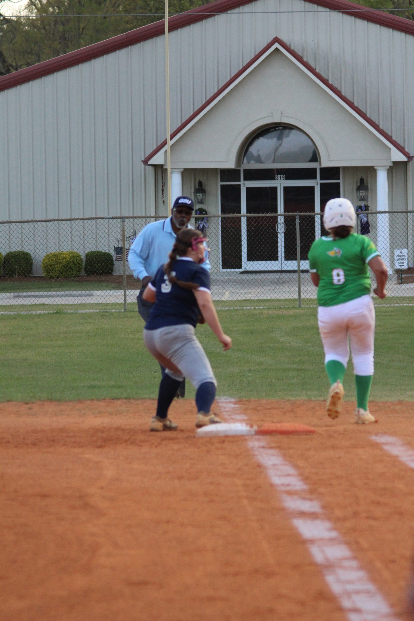 A softball player in green runs to first base as a player in navy prepares to catch, with an umpire observing closely.