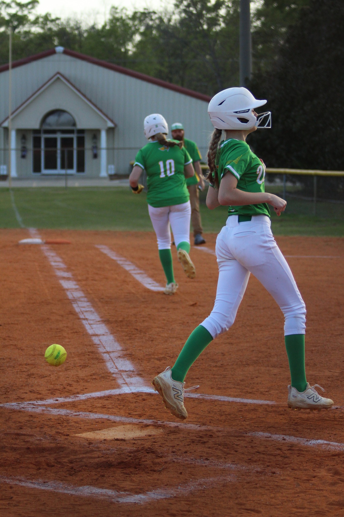 Two female softball players in green uniforms on a dirt field, one crossing home plate as a yellow softball lies near the base line.
