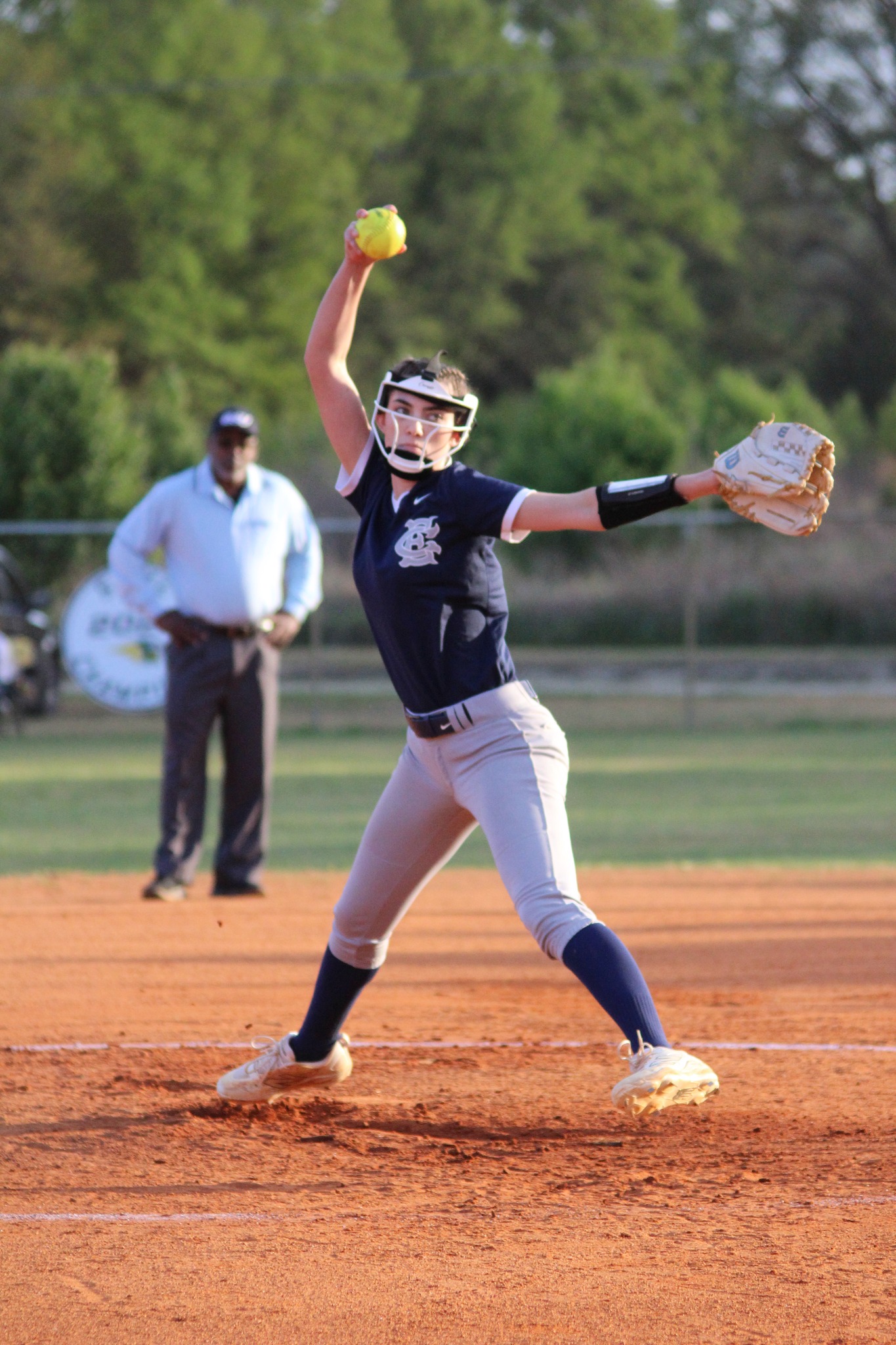 Softball pitcher in navy and gray uniform preparing to throw the ball, with an umpire and trees in the background on a sunny day.