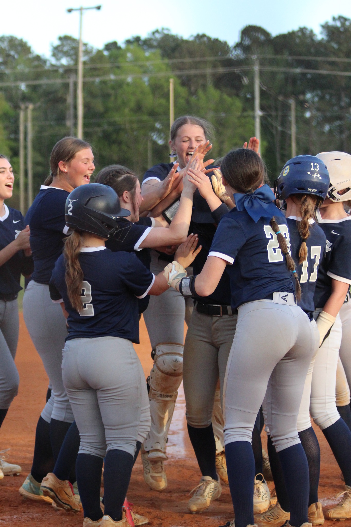 A group of softball players in navy jerseys celebrate on a baseball field, smiling and gathering around a teammate, with trees visible in the background.