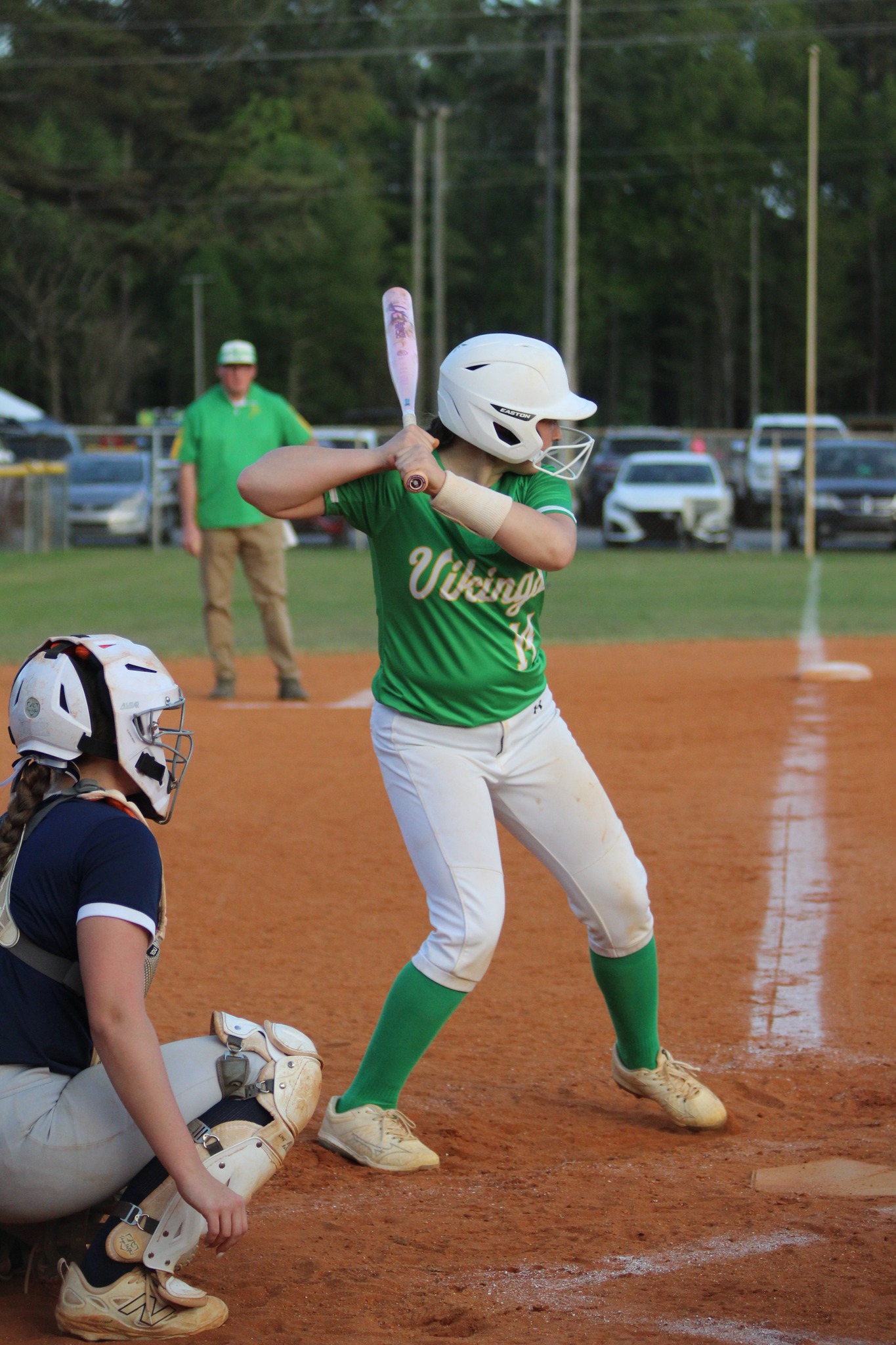 A softball player in a green jersey prepares to bat while a catcher crouches behind her on a dirt field.