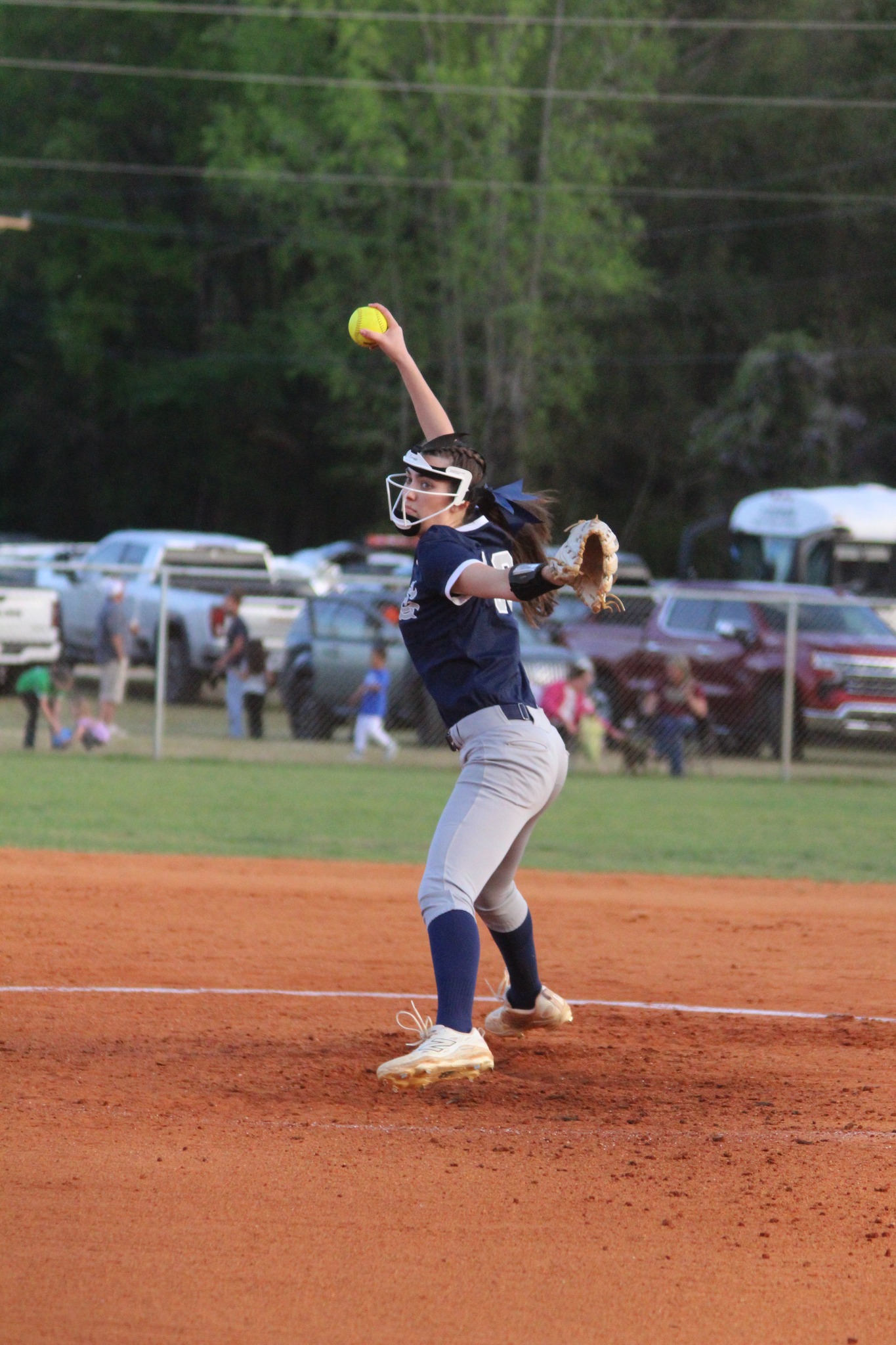 A softball player in a navy jersey and gray pants pitches the ball on a dirt field, with cars and spectators in the background.