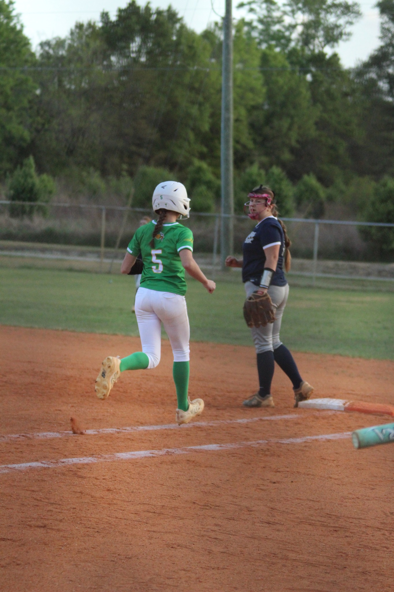 A softball player in green runs toward home plate as a catcher in navy blue and white stands nearby on a dirt field.