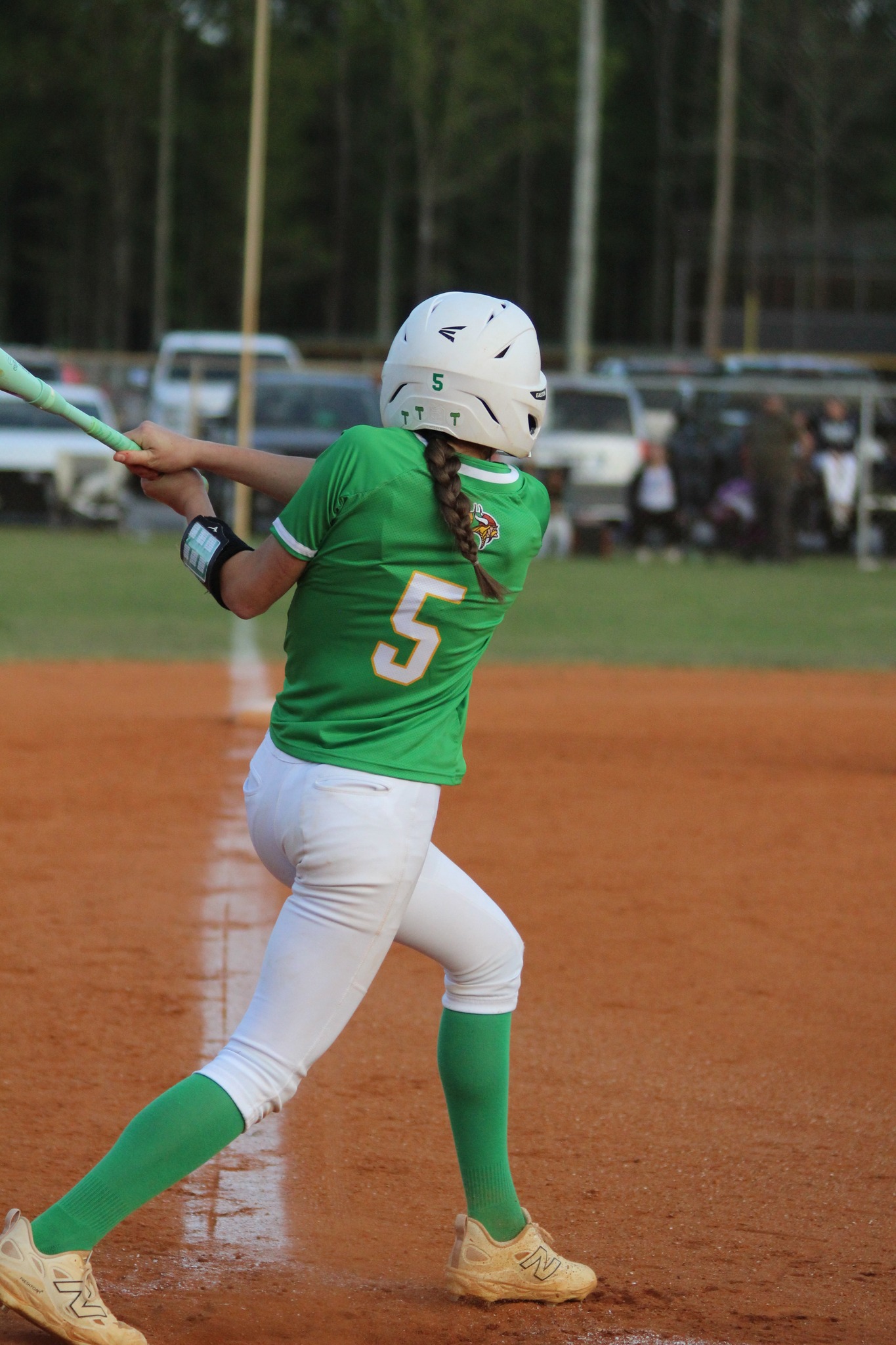 A softball player wearing a green jersey with number 5 swings a bat on a dirt field, with cars and spectators in the background.