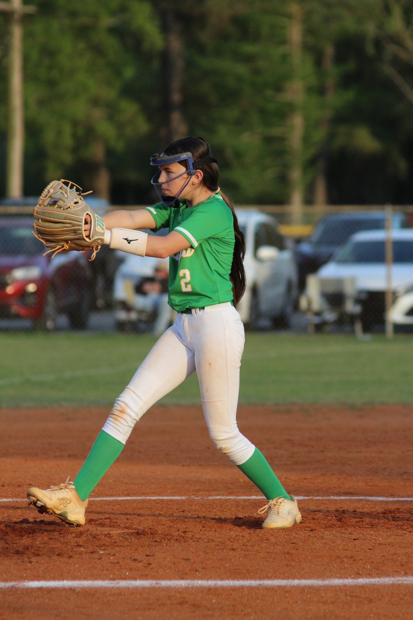 A softball player in a green jersey and white pants is pitching on a dirt field, with cars and trees in the background.