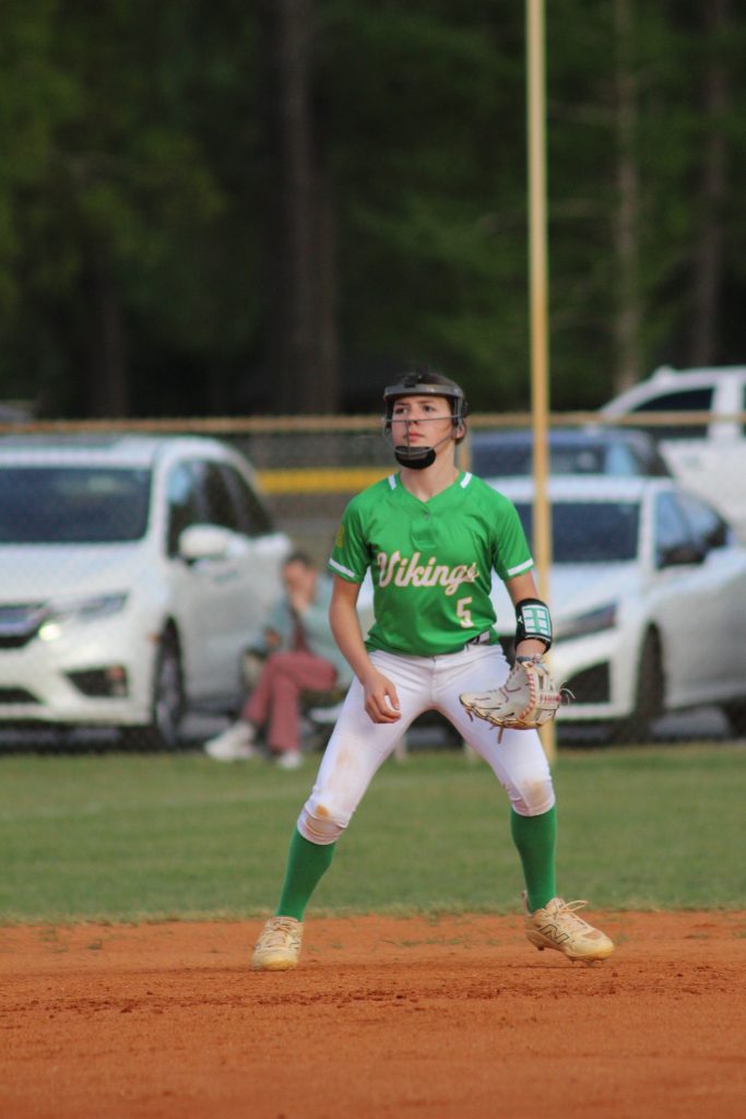 Baseball player in green jersey and white pants stands ready on the field, with cars and a person sitting in the background.