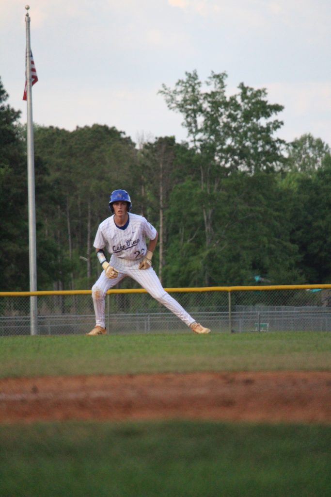 A baseball player in white uniform and blue helmet leads off a base on a field, with trees and a flagpole in the background.