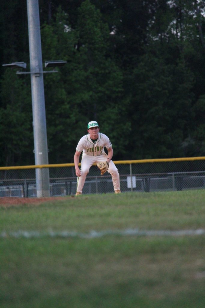 Baseball player in a white uniform and helmet stands ready in the infield during a game, with trees and a fence in the background.