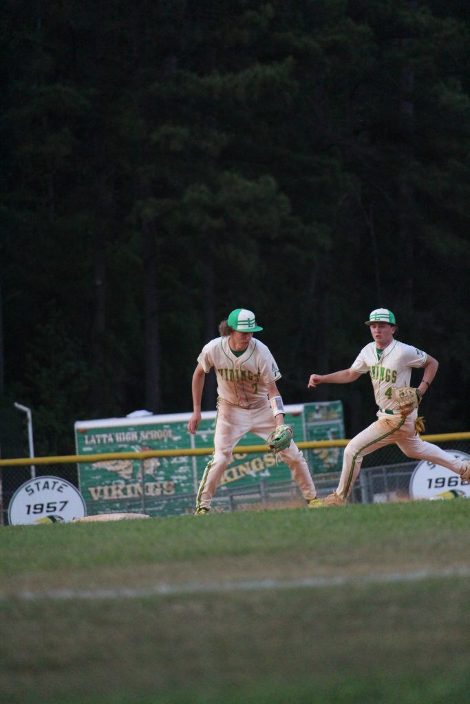 Two high school baseball players in uniforms and green hats field a play on a baseball diamond with a "Vikings" team banner in the background.