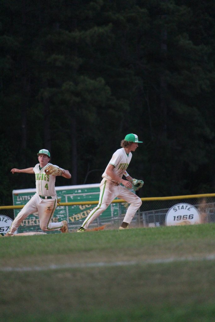 Two baseball players in white and green uniforms play on a field, one running and the other preparing to throw, with trees and signs in the background.
