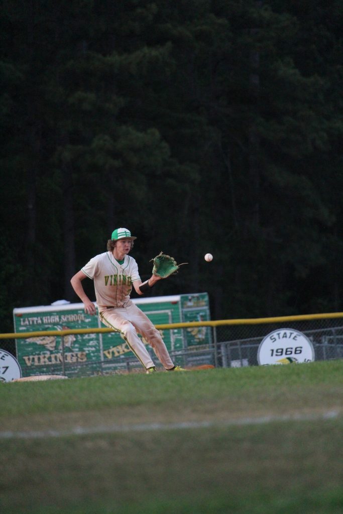 A baseball player in a green and white uniform attempts to catch a ball mid-air on a field, with trees and banners in the background.