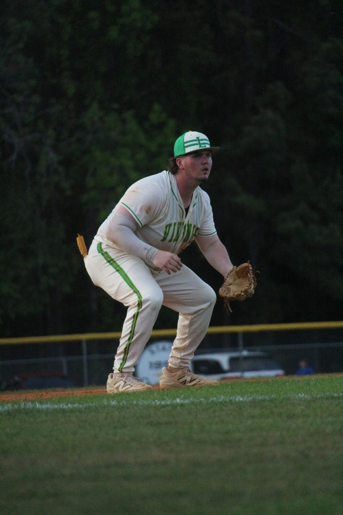 Baseball player in white and green uniform crouching on the field, ready in a defensive position, with glove on left hand and trees in the background.