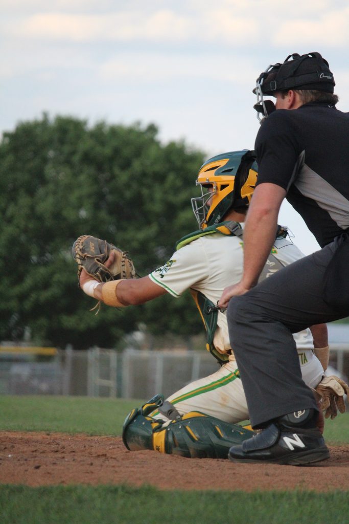 A baseball catcher in green and yellow gear is kneeling, glove extended, with an umpire in black crouched behind during a game outdoors.