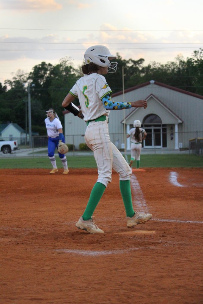A softball player in a white and green uniform stands on a dirt field, preparing to bat, with other players and a building in the background.