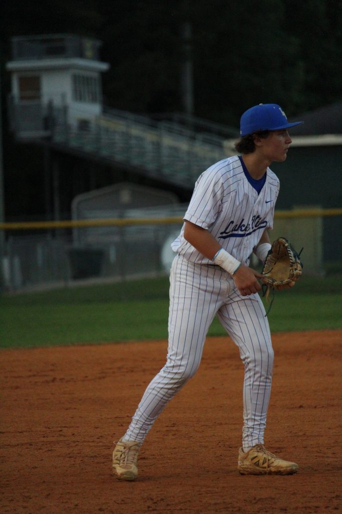 A baseball player in a white pinstripe uniform and blue cap stands ready on an infield dirt field, holding a glove.