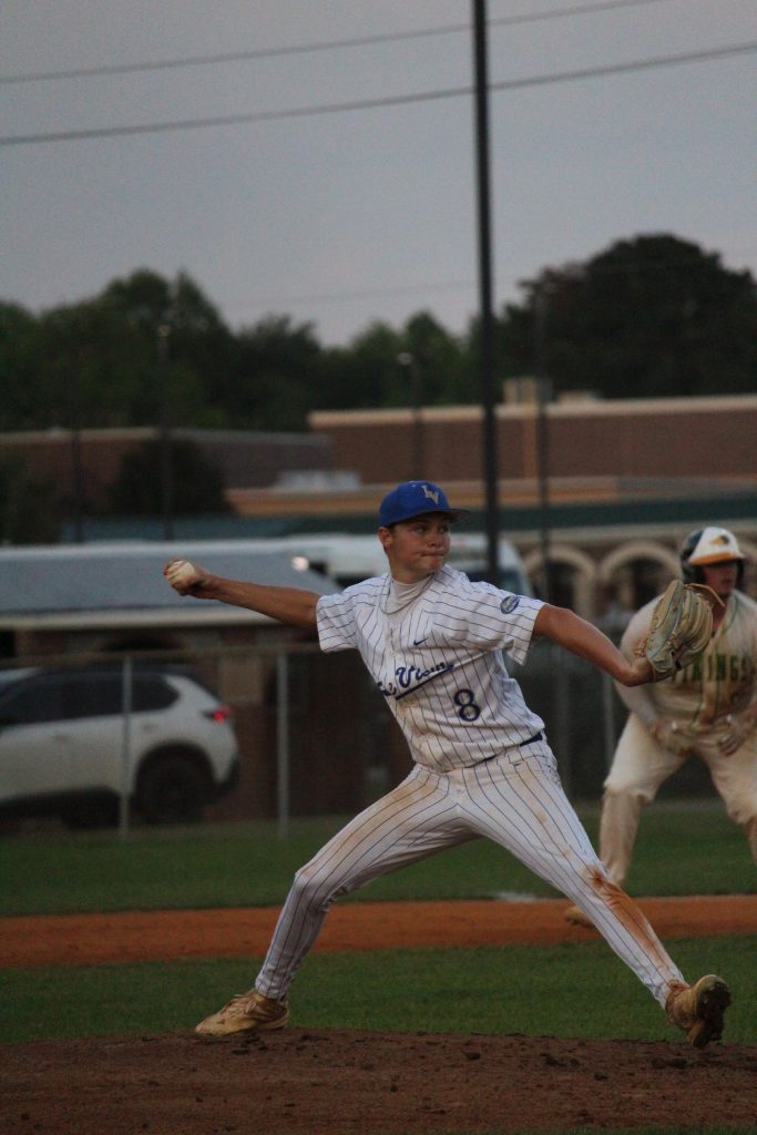 A baseball pitcher in a white pinstripe uniform winds up to throw a pitch during a game, with another player in the background.