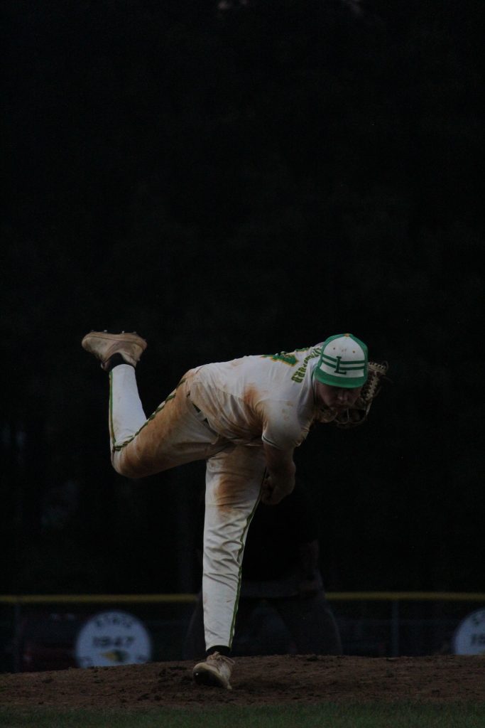 A baseball pitcher in a white and green uniform throws a pitch on a dirt mound, with a dark background behind him.