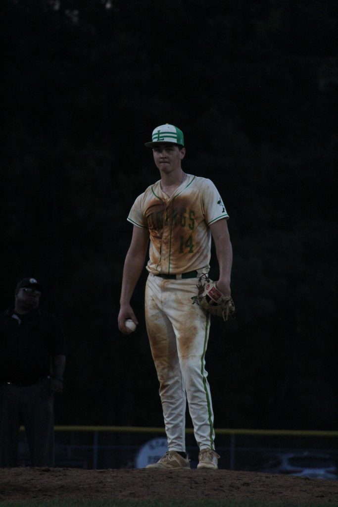A baseball player in a dirty white and green uniform stands on the mound, holding a ball and glove, with an umpire in the background.