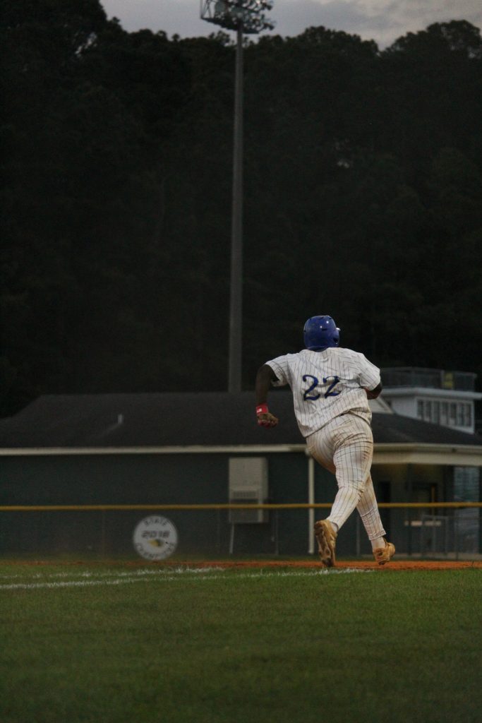 Baseball player wearing number 22 runs on a base path during a game at dusk, with trees and a building in the background.