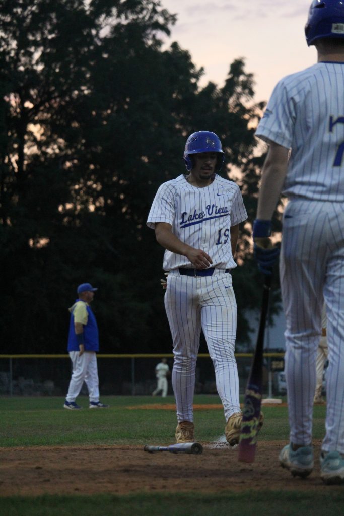 A baseball player wearing a "Lake View" uniform walks past home plate at dusk, with teammates and a coach visible in the background.