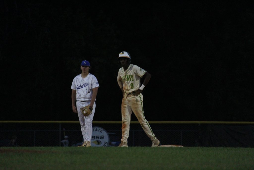 Two baseball players stand on the field at night, one on a base in a dirt-stained uniform, another nearby in a striped white uniform.