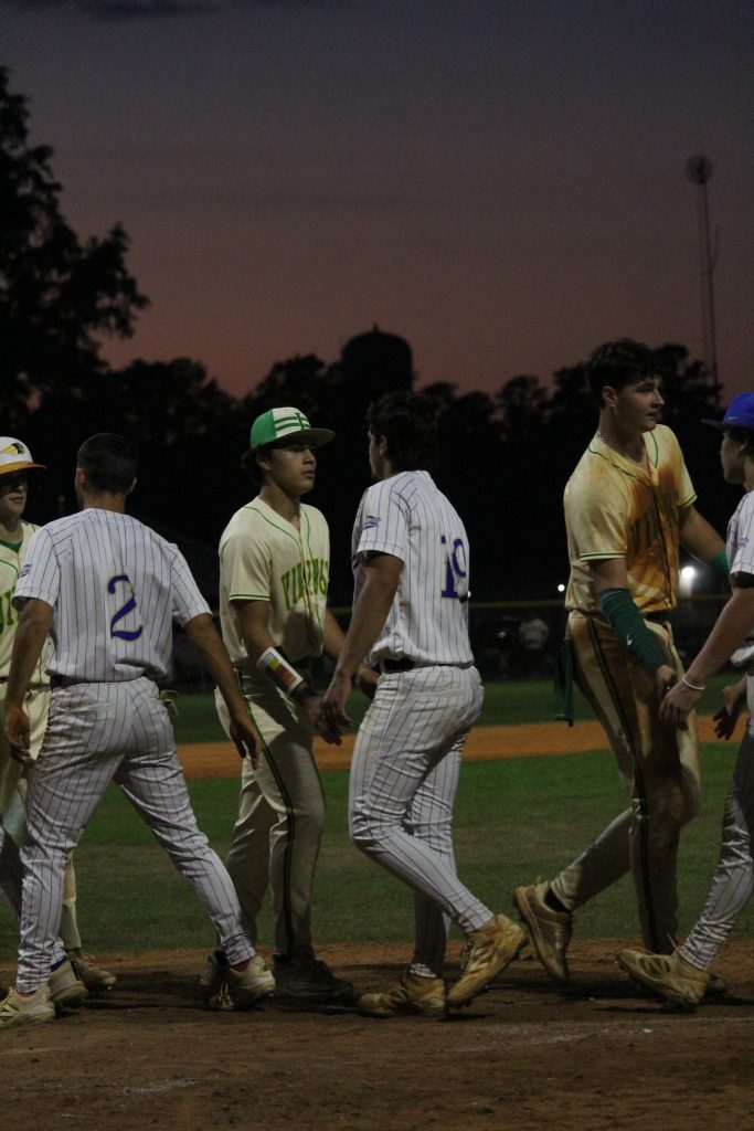 Baseball players from two teams shake hands on the field after a game at dusk, with trees and a water tower in the background.