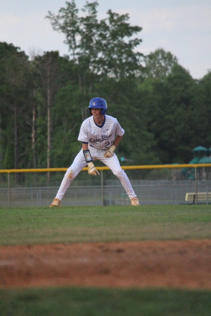 A baseball player in a white uniform and blue helmet stands ready on a grassy field, preparing to run. Trees and a yellow fence are in the background.