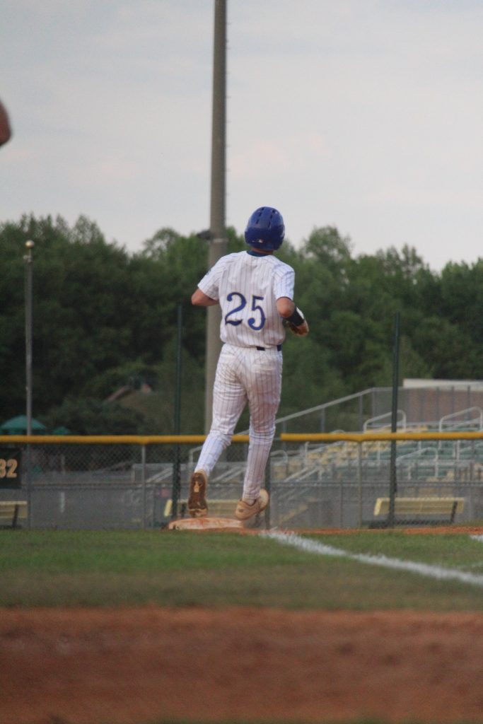 A baseball player wearing a blue helmet and jersey number 25 runs past a base on an outdoor baseball field.