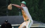 A baseball pitcher in a white and green uniform winds up to throw, with an umpire and trees in the background.