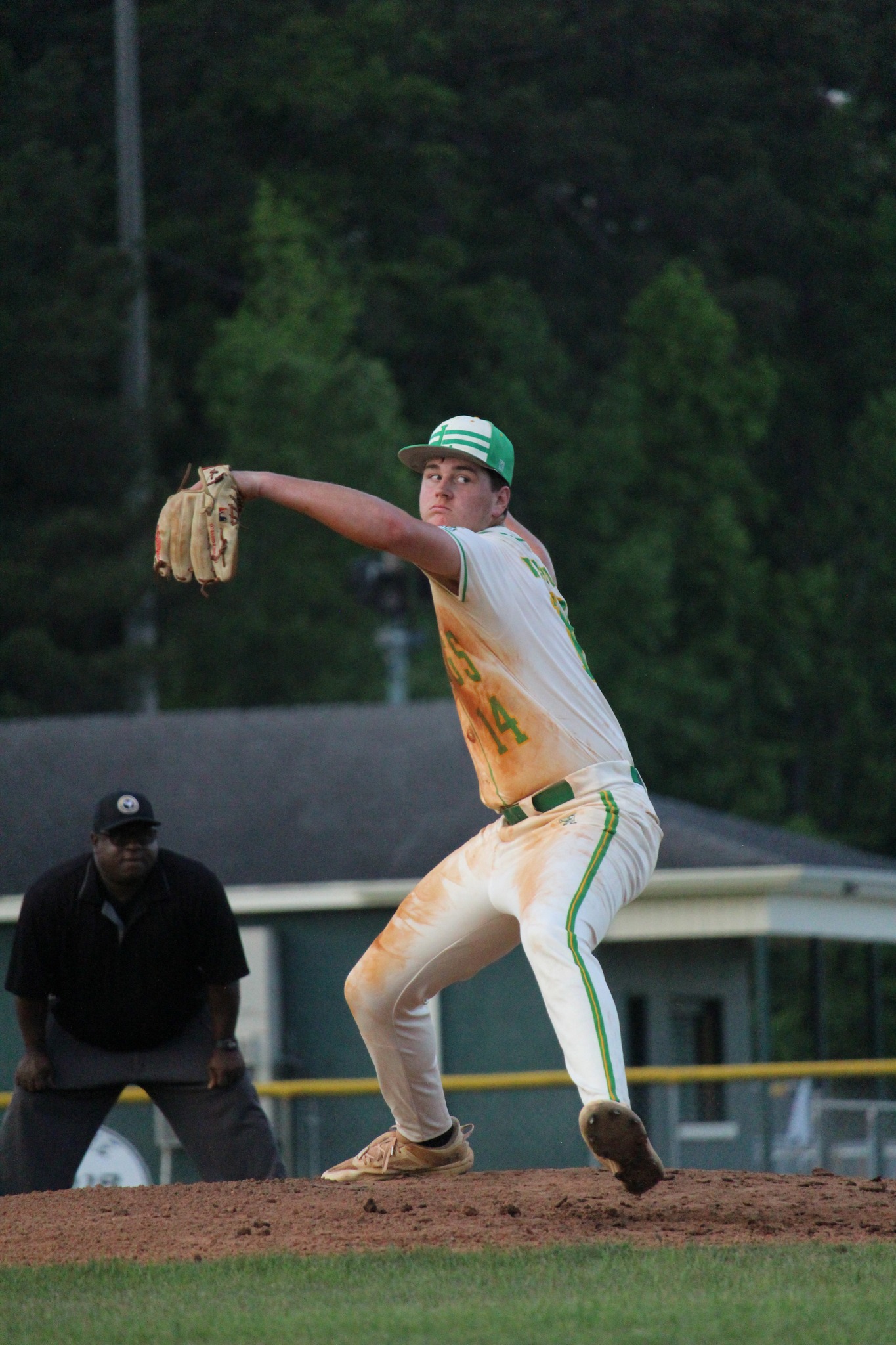 A baseball pitcher in a white and green uniform winds up to throw, with an umpire and trees in the background.
