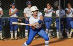 A softball player at bat readies to swing, while teammates in blue and white uniforms watch from behind the fence.