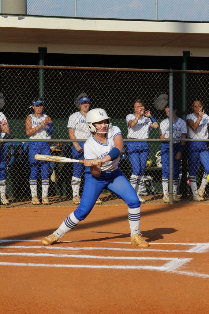 A softball player at bat readies to swing, while teammates in blue and white uniforms watch from behind the fence.