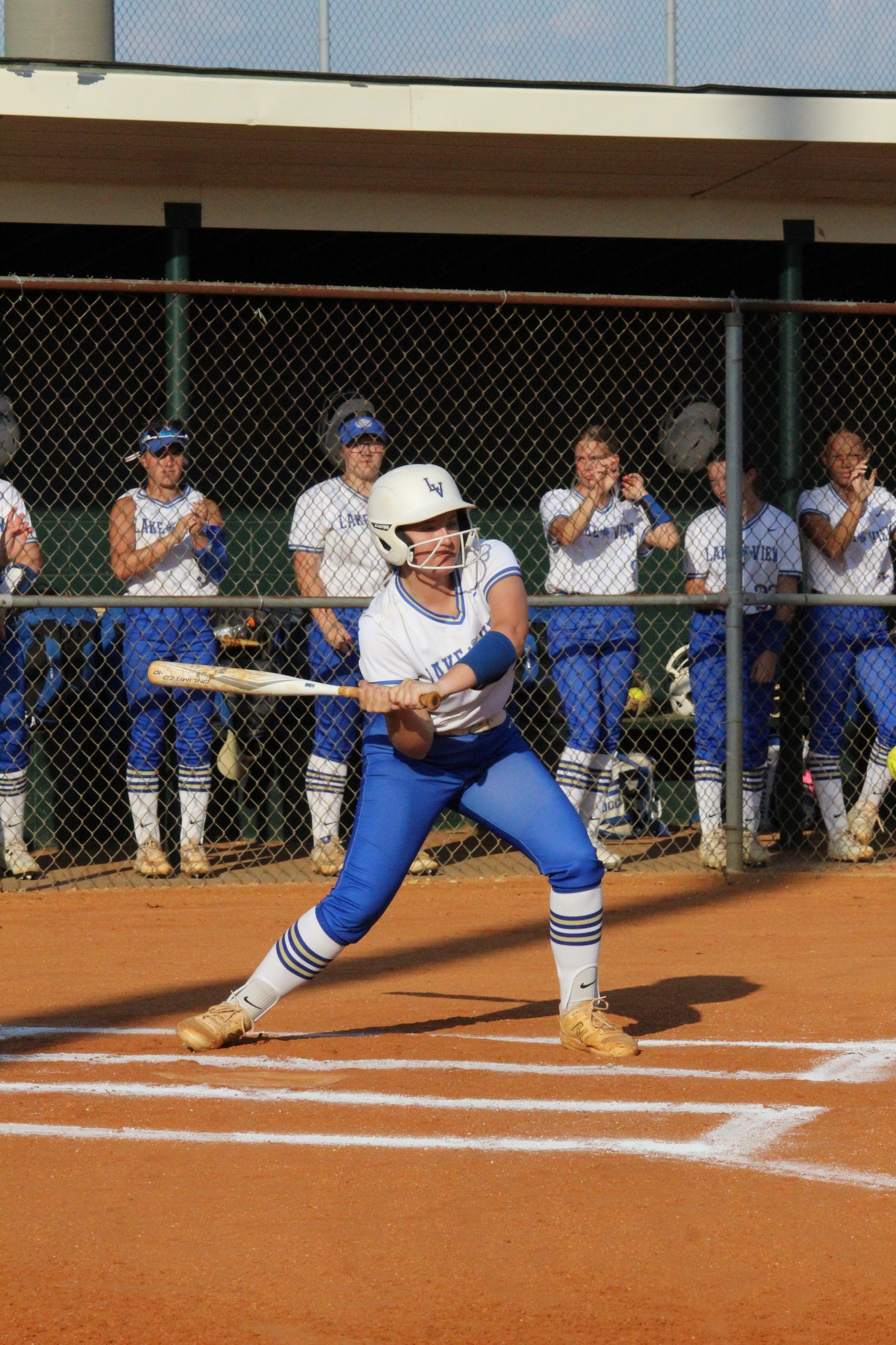A softball player at bat readies to swing, while teammates in blue and white uniforms watch from behind the fence.