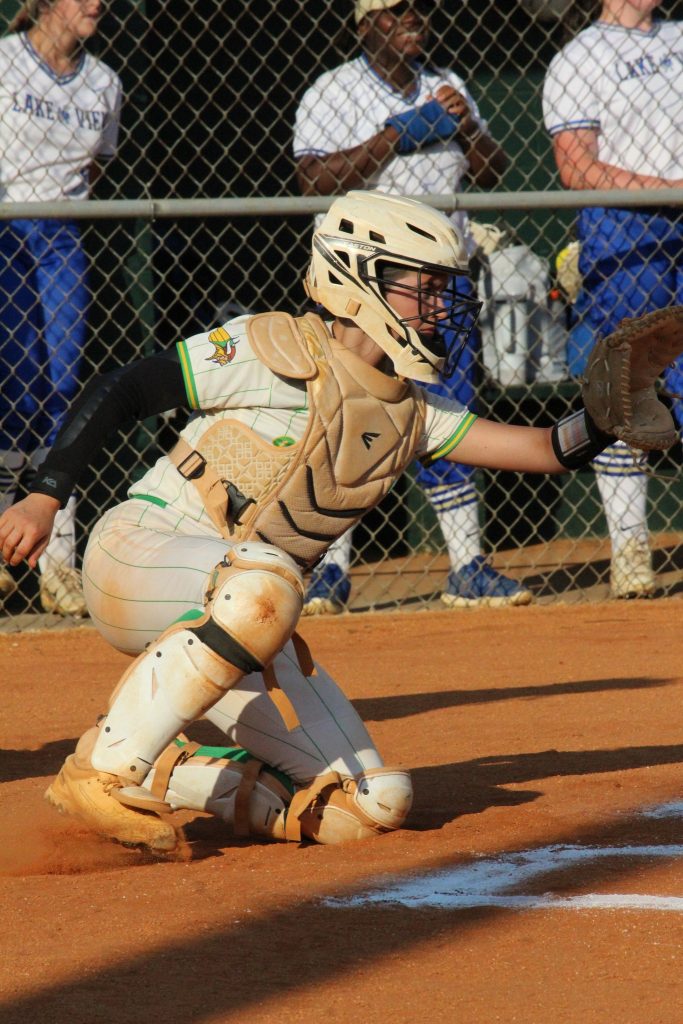 A baseball catcher in full gear crouches behind home plate, reaching out with a glove to catch a pitch during a game.