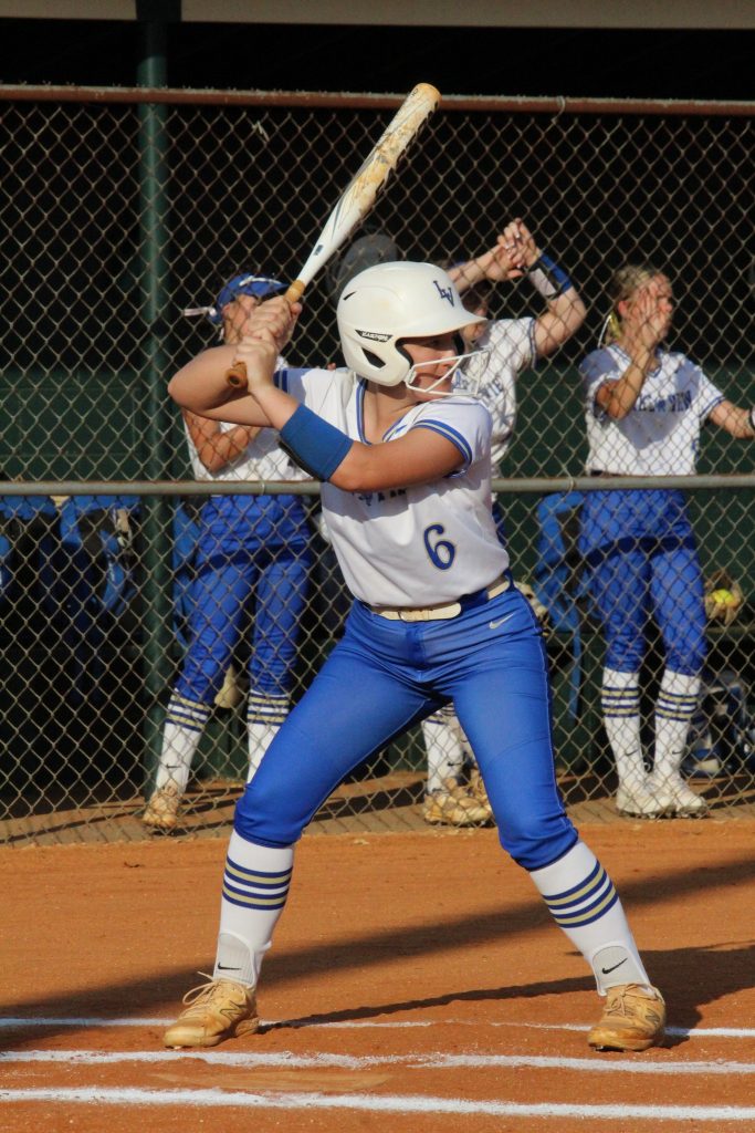 A softball player in blue and white uniform prepares to bat, while teammates watch from behind a fence on the field.