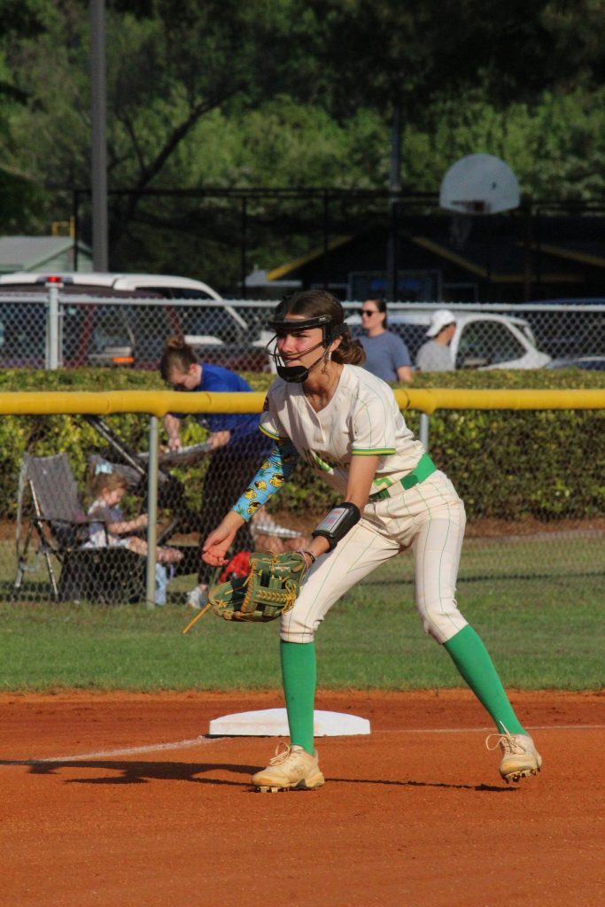 A softball player in a white uniform with green socks stands ready on the field near a base, with spectators and cars in the background.