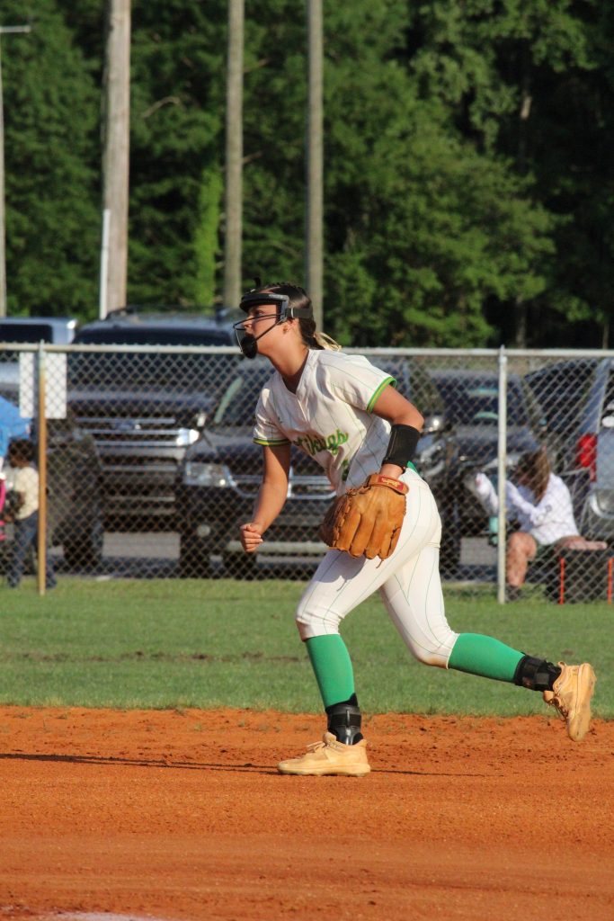 A softball player in uniform prepares to throw the ball on a dirt field, with cars and spectators visible in the background.