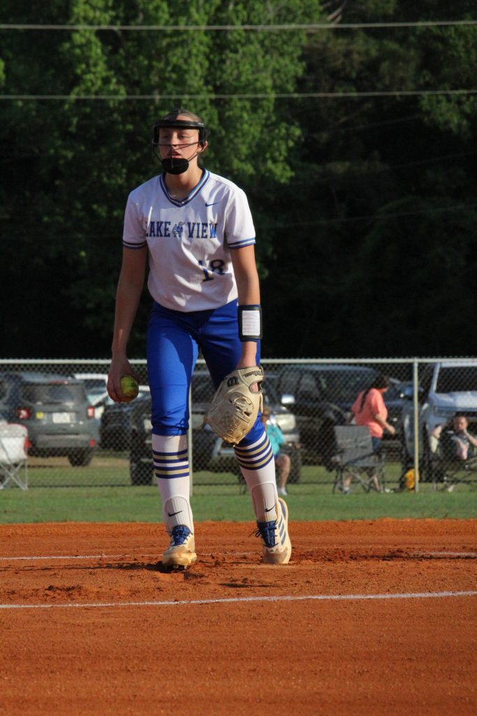 A softball player wearing a white and blue uniform stands on a field, preparing for action, with spectators and cars visible in the background.