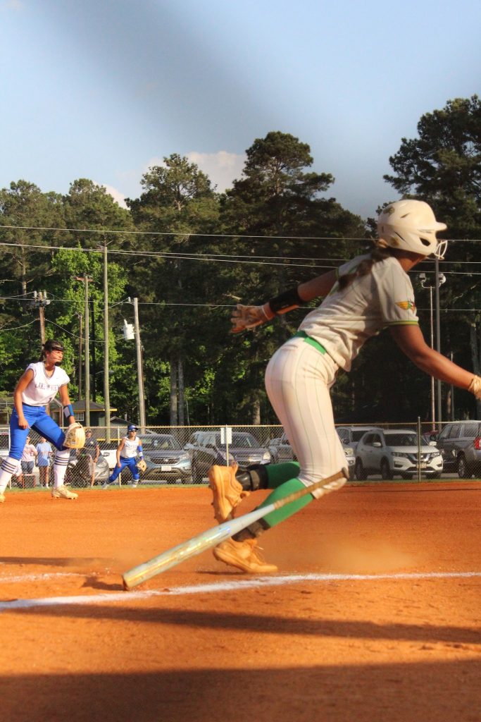 A softball player runs toward first base as an opposing player prepares to catch the ball on a sunny outdoor field.