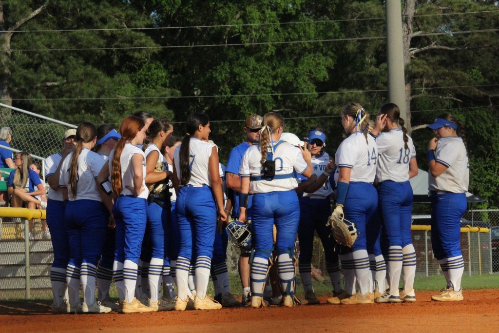 A softball team in white and blue uniforms gathers in a huddle on the field, surrounded by trees and a fence in the background.