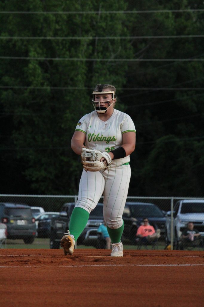 A softball player in a white and green uniform stands on the dirt field, wearing a helmet and glove, with cars and trees in the background.