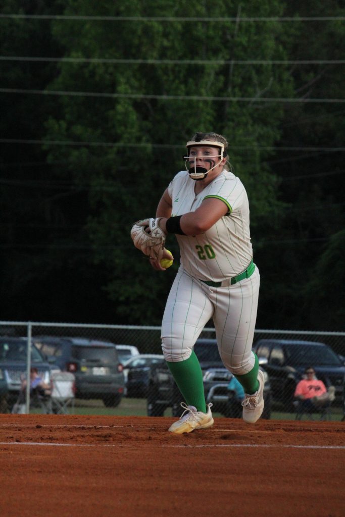 A softball player in white and green uniform prepares to throw the ball on a field, with cars and trees in the background.